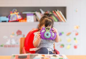 Little girl peeking on a toy camera inside a preschool classroom